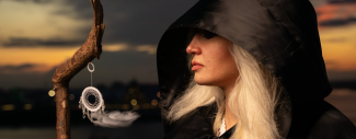 photo d'une femme blonde encapuchonnée de noir et portant un bâton avec un attrape-rêves